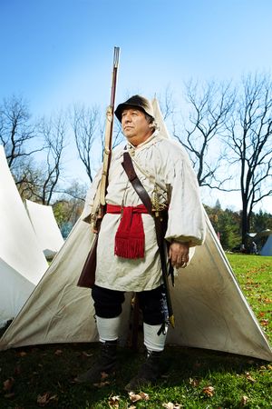 Colonial soldier in period costume holding musket rifle near white tent outdoors.