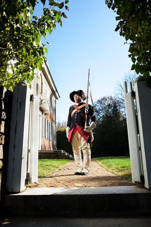 Black soldier in Revolutionary War uniform carrying a musket walks through a gate at a historic colonial site.