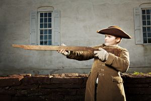 Young boy in colonial tricorn hat aims a musket during an American Revolutionary War reenactment.