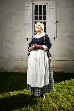 Woman in 18th century colonial servant costume with white apron and bonnet stands outside historic building.