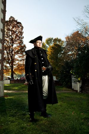 Man in black colonial officer coat and tricorn hat stands on grass at a historic autumn estate.