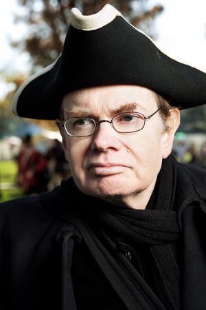 Man wearing black tricorn hat and scarf portrays a colonial era character at a historical reenactment.