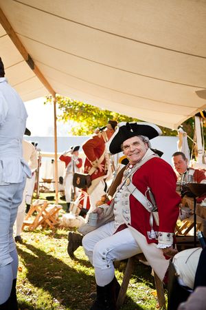 Smiling man in British Redcoat uniform sits under a tent during an American Revolutionary War reenactment.