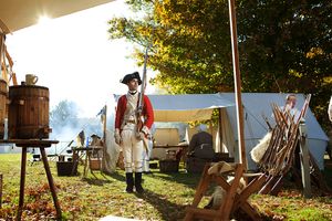 Soldier in British Redcoat uniform stands in a Revolutionary War reenactment camp with tents and equipment.