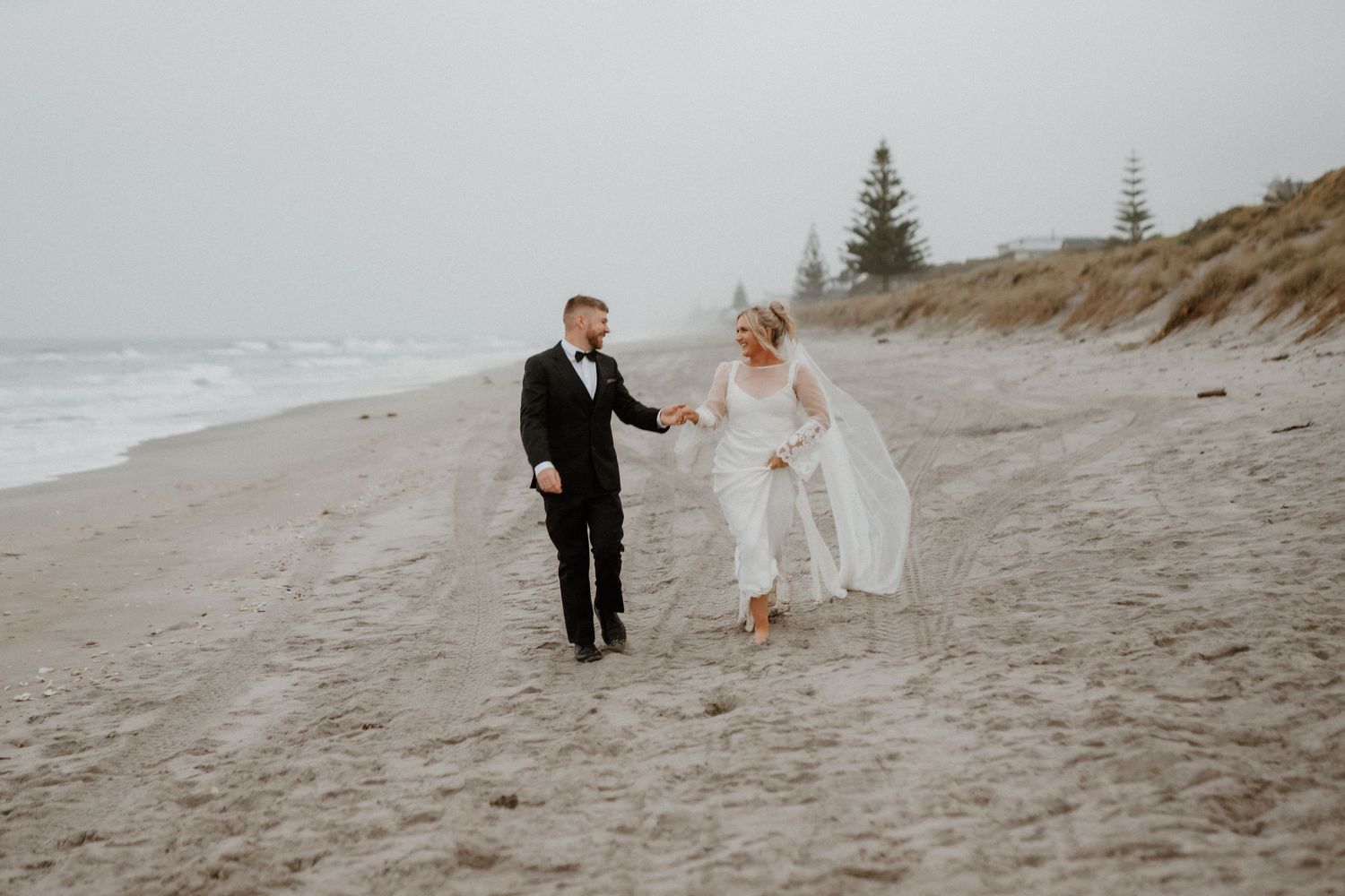 Newlyweds joyfully walk together on a misty beach with evergreen trees in the background.