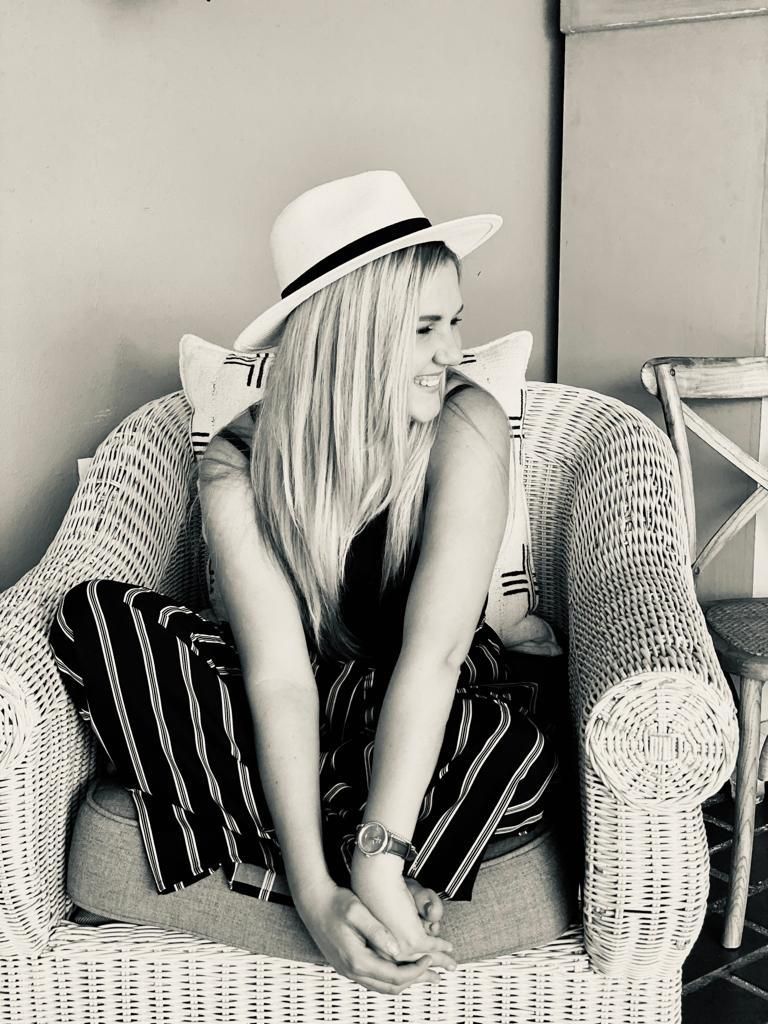 Blonde woman in a hat sitting in a wicker chair smiling outdoors.