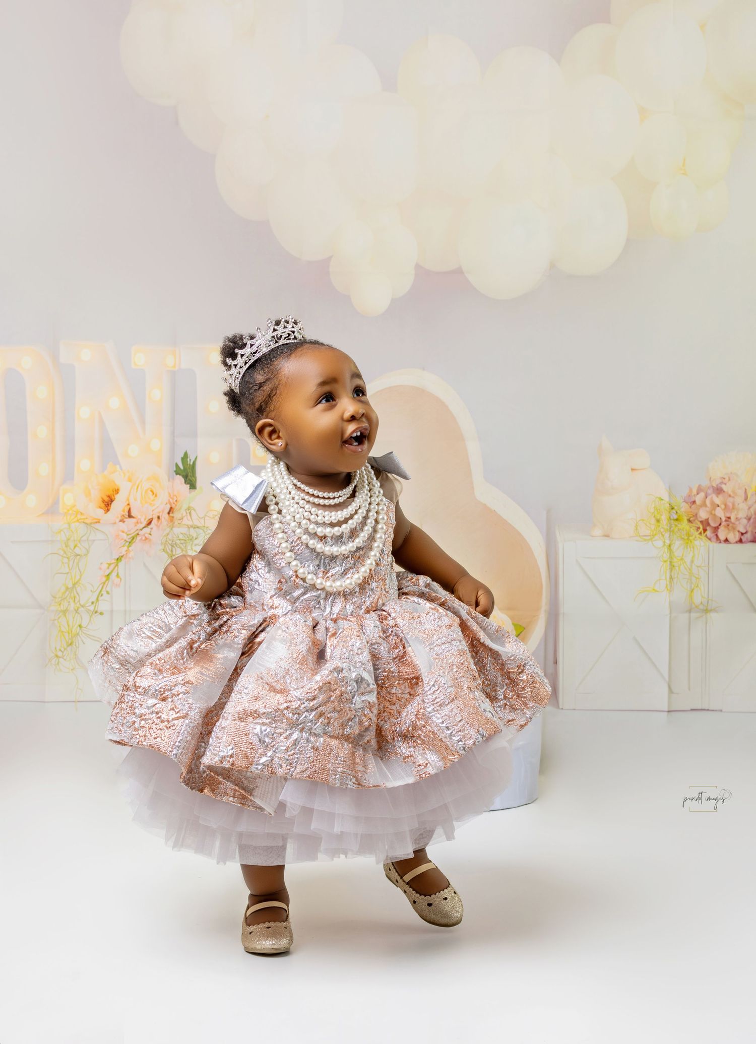 Baby in sparkly dress and pearls twirling during birthday photoshoot with soft backdrop.