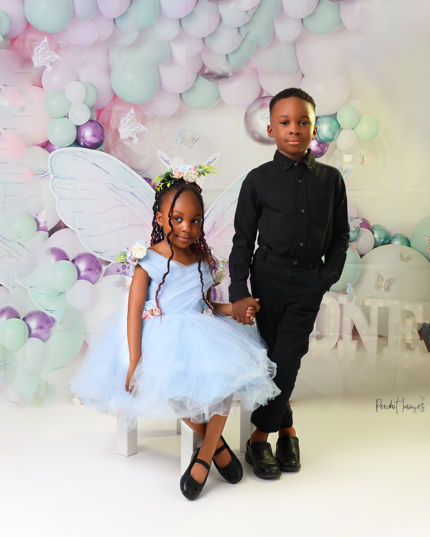 Two children pose in formal attire against pastel balloon arch backdrop.
