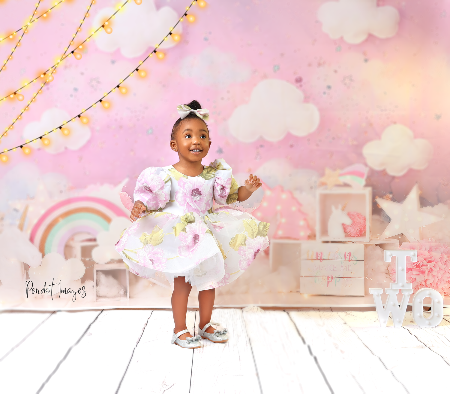 Toddler in white dress poses against dreamy pink backdrop with clouds and string lights.