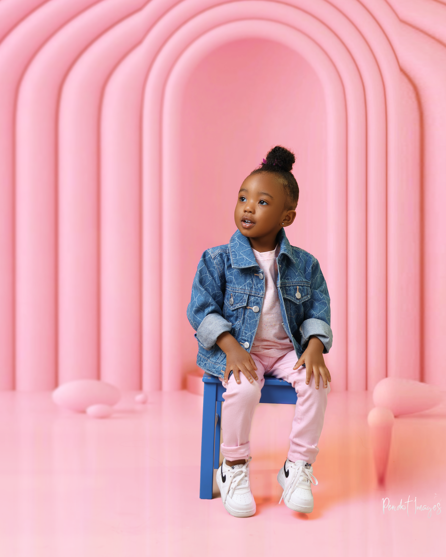 Child in denim jacket poses against pink arched backdrop.