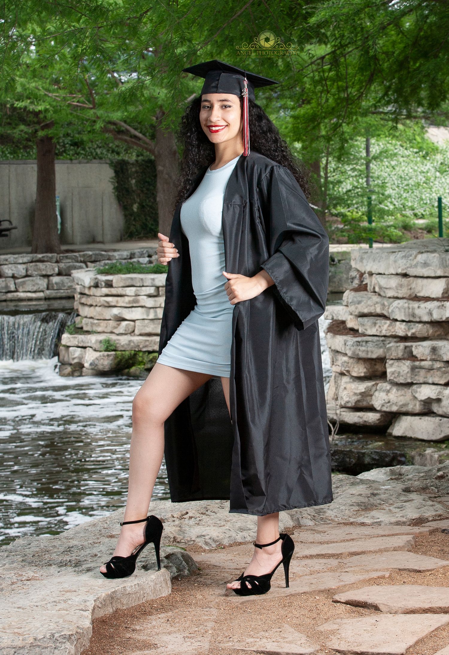 Graduate in black cap and gown posing by stone waterfall.