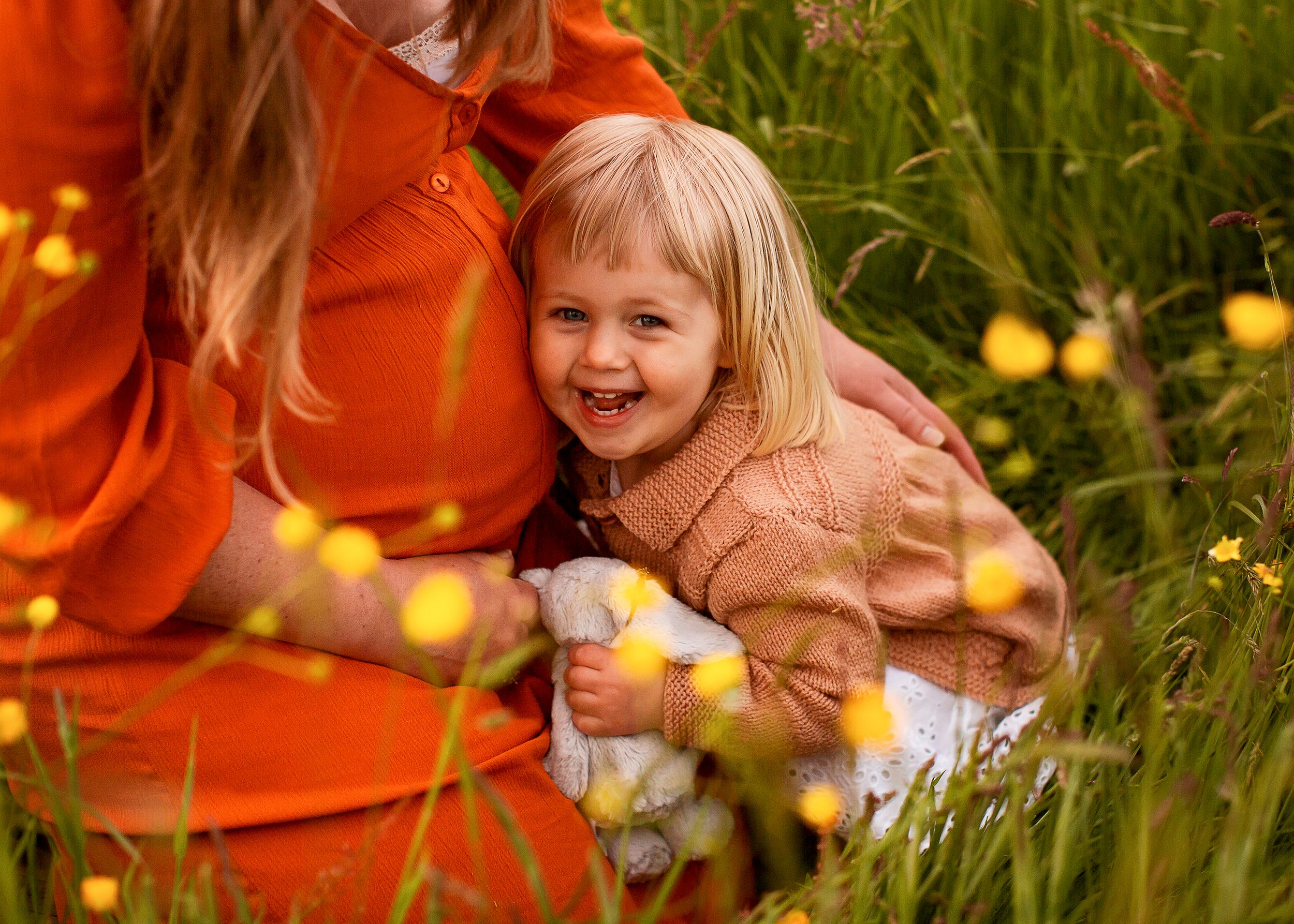 Happy smiling toddler cuddles her mum's pregant bump in wildflower meadow photoshoot