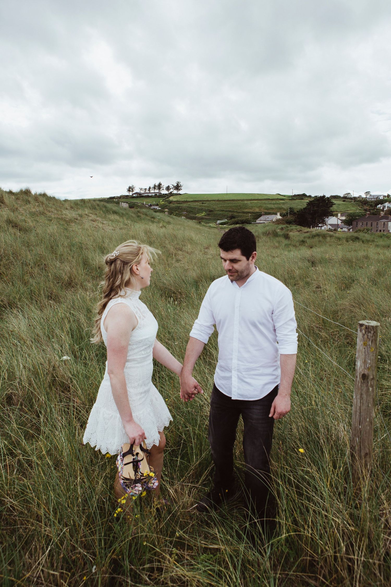 A couple in casual wedding attire stands together in a grassy field on a cloudy day.