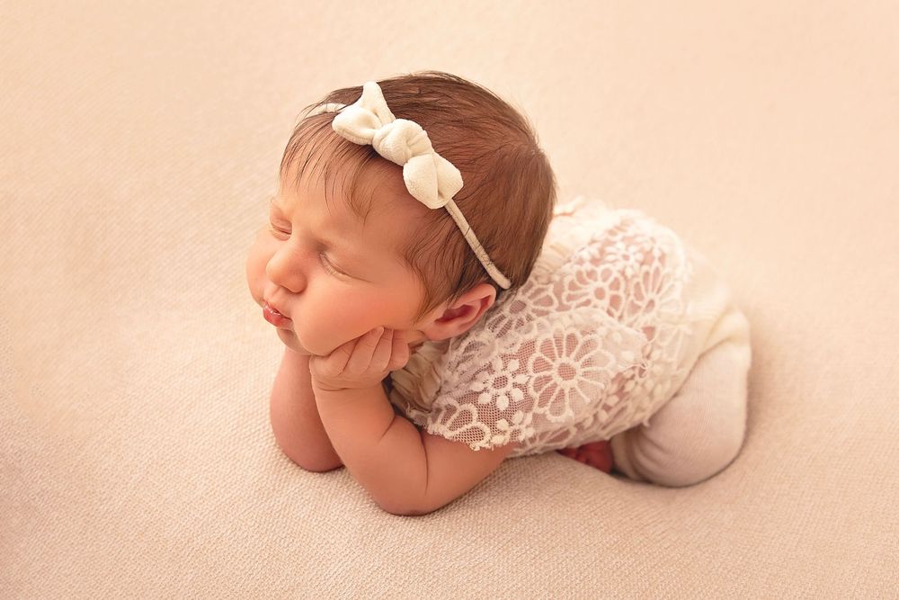 Sleeping newborn baby girl in lace romper with bow headband in froggy pose.