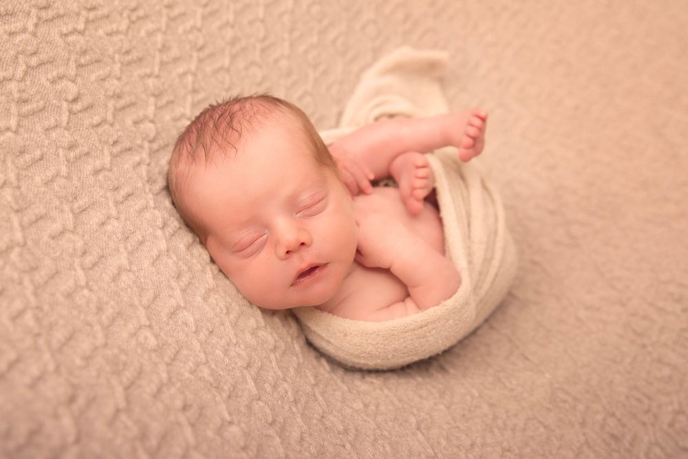 Peacefully sleeping newborn wrapped in white swaddle curled on textured beige blanket.