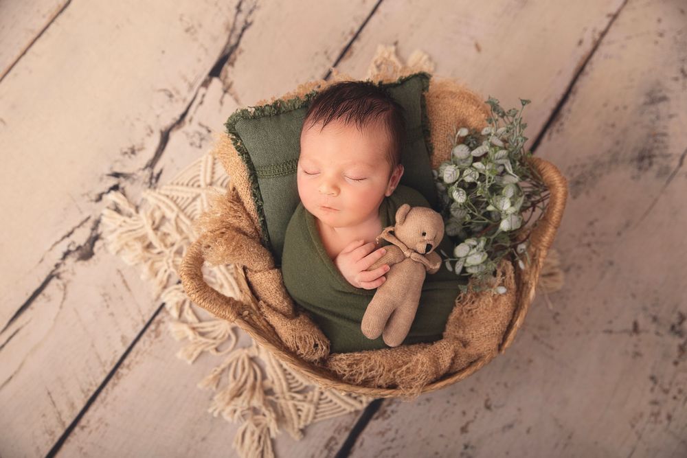 Sleeping newborn in green wrap nestled in basket with teddy bear and flowers.