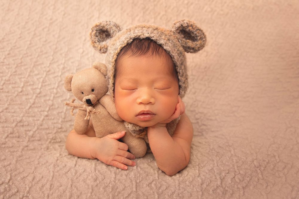 Sleeping newborn wearing bear ear bonnet cuddling matching teddy bear on beige blanket.