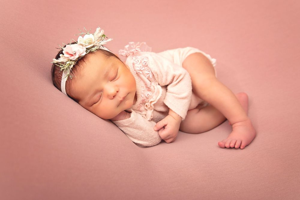 Sleeping newborn girl in pink lace outfit with floral headband on pink backdrop.