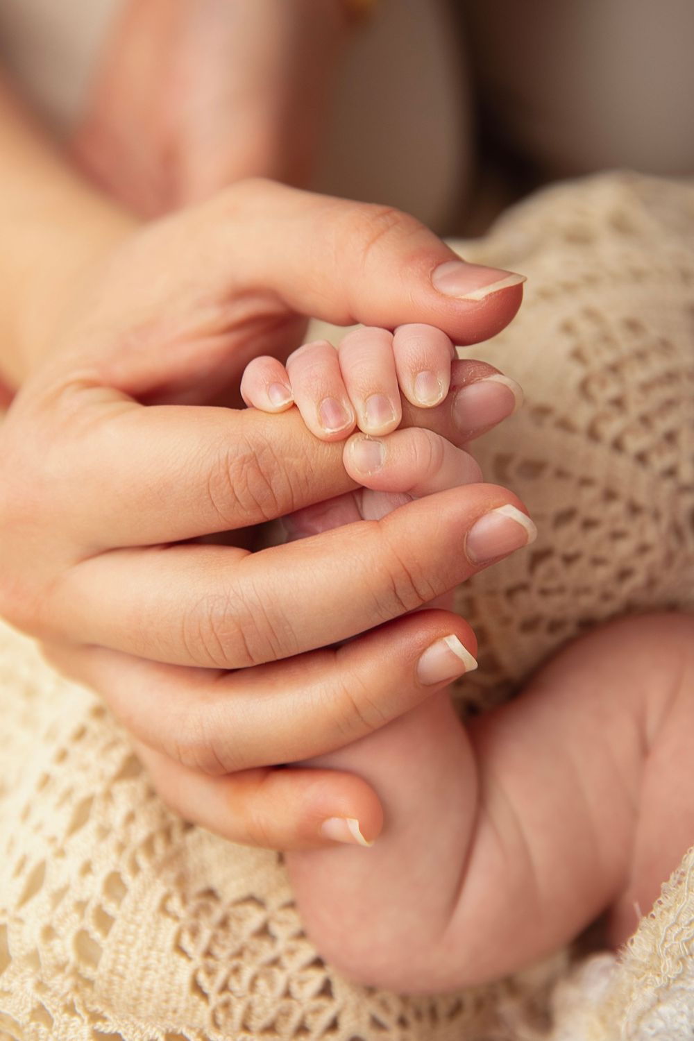 Adult hands gently cradling tiny newborn fingers wrapped in cream knit blanket.