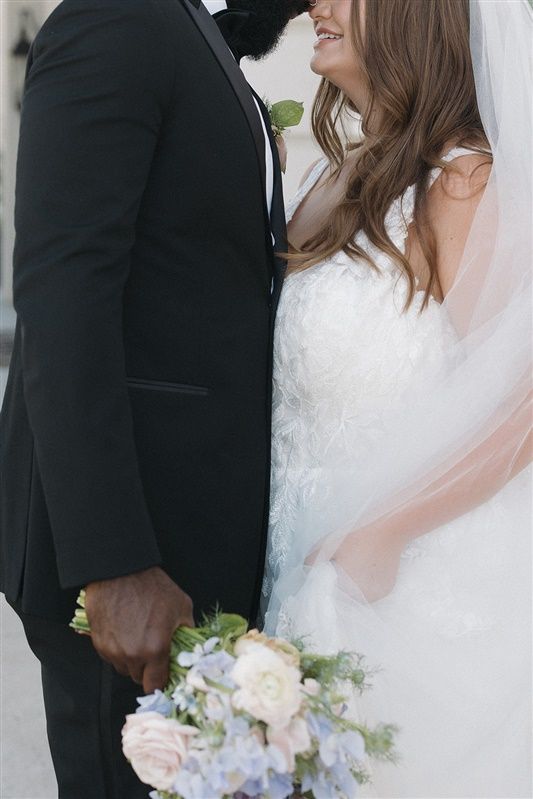 Newlywed couple in wedding attire standing close together with pastel bouquet of pink and blue flowers.