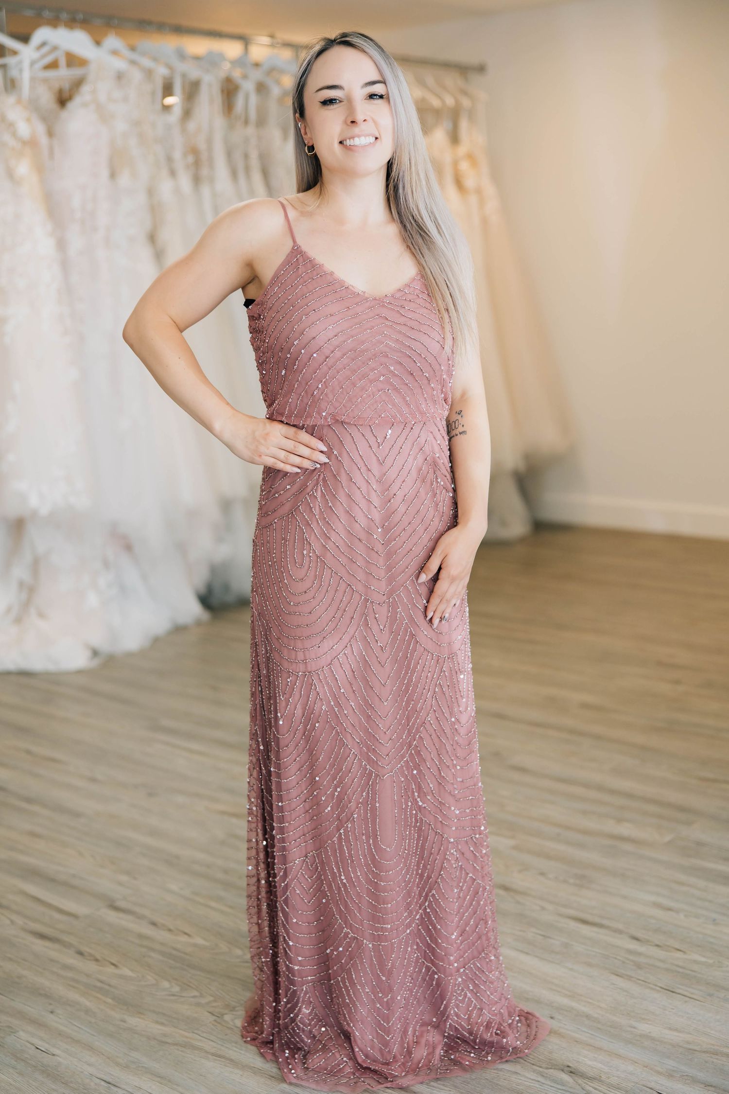 Smiling woman in mauve beaded spaghetti strap maxi gown posing in bridal boutique.