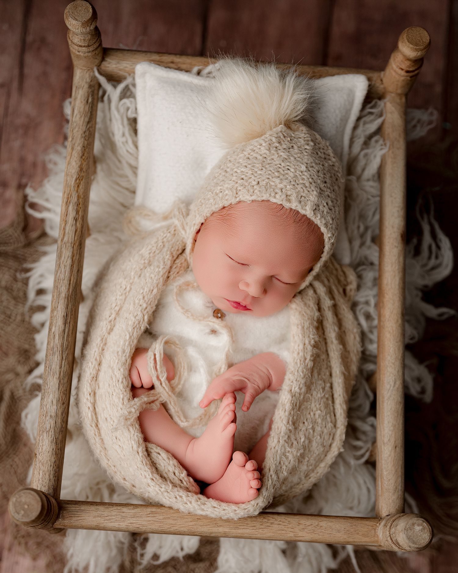 A sleeping newborn wrapped in a cream-colored lace wrap lies peacefully in a rustic wooden bed prop.