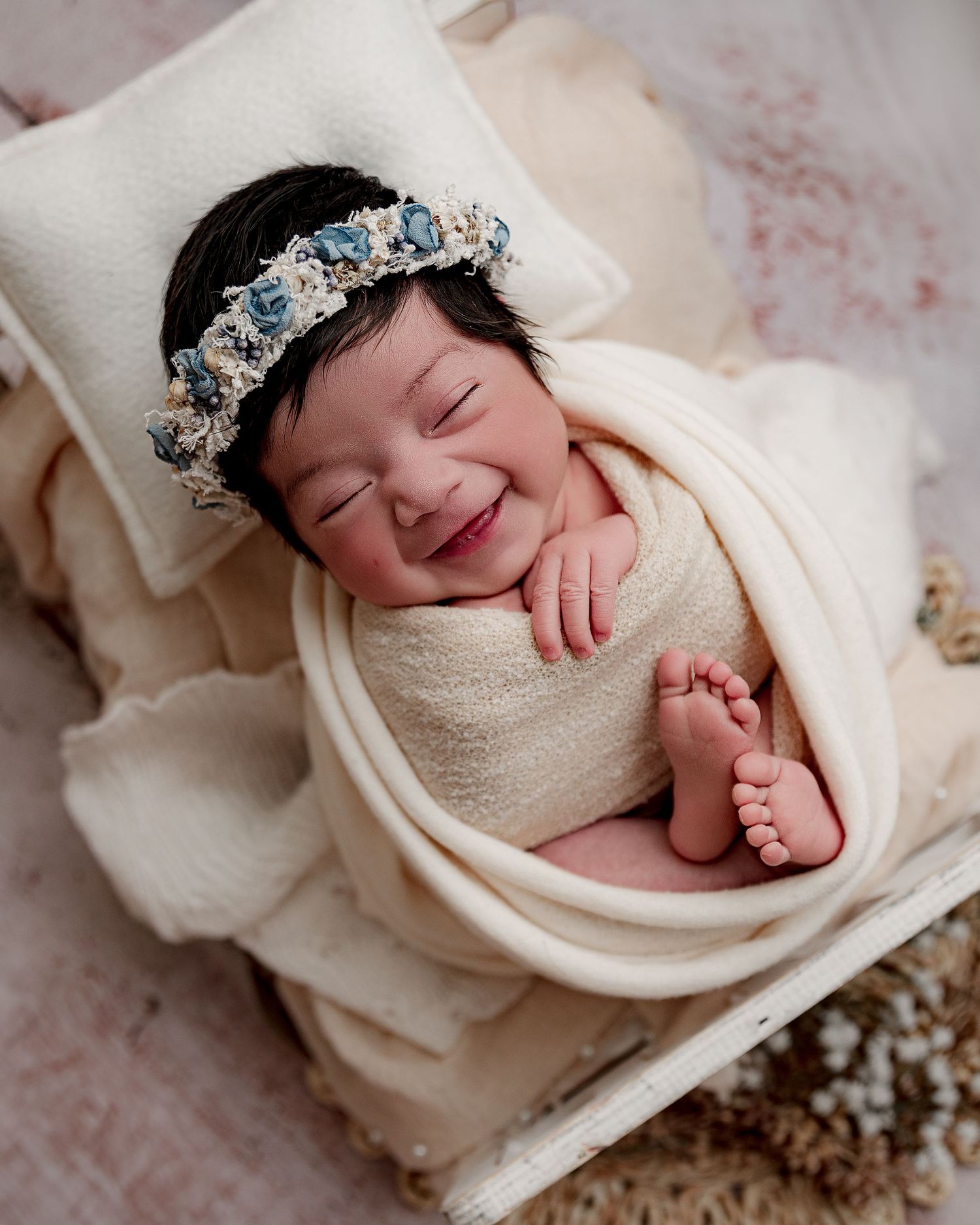 Smiling newborn wearing blue crown wrapped in cream fabric on white surface.