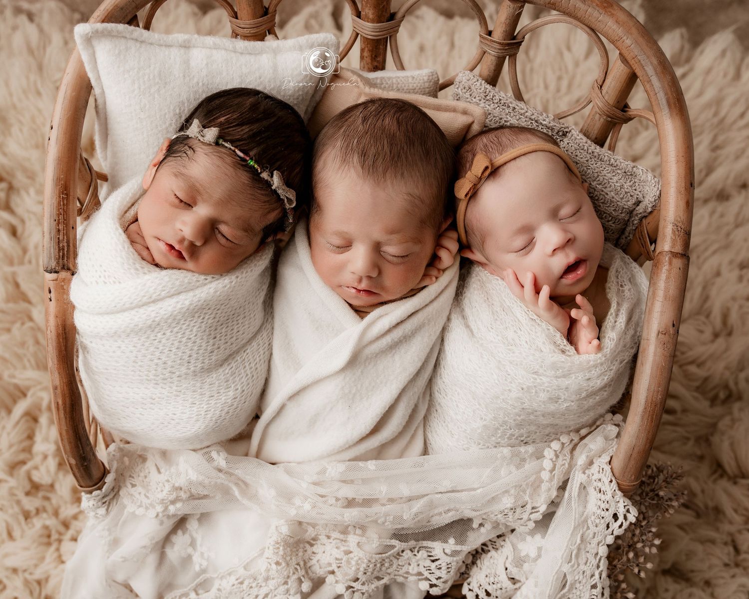 Three sleeping newborns swaddled in white blankets lie side by side in a woven basket on lace fabric.