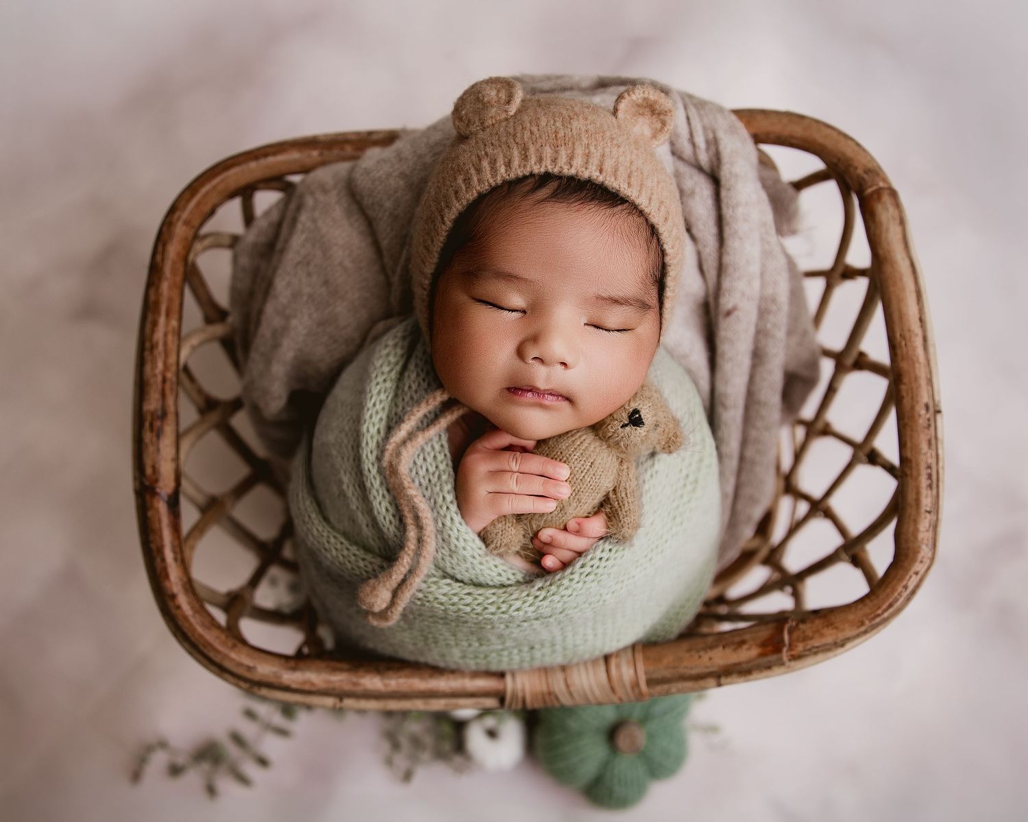 Cozy sleeping newborn in knit bonnet and wrap nestled in woven basket with soft decorative elements.