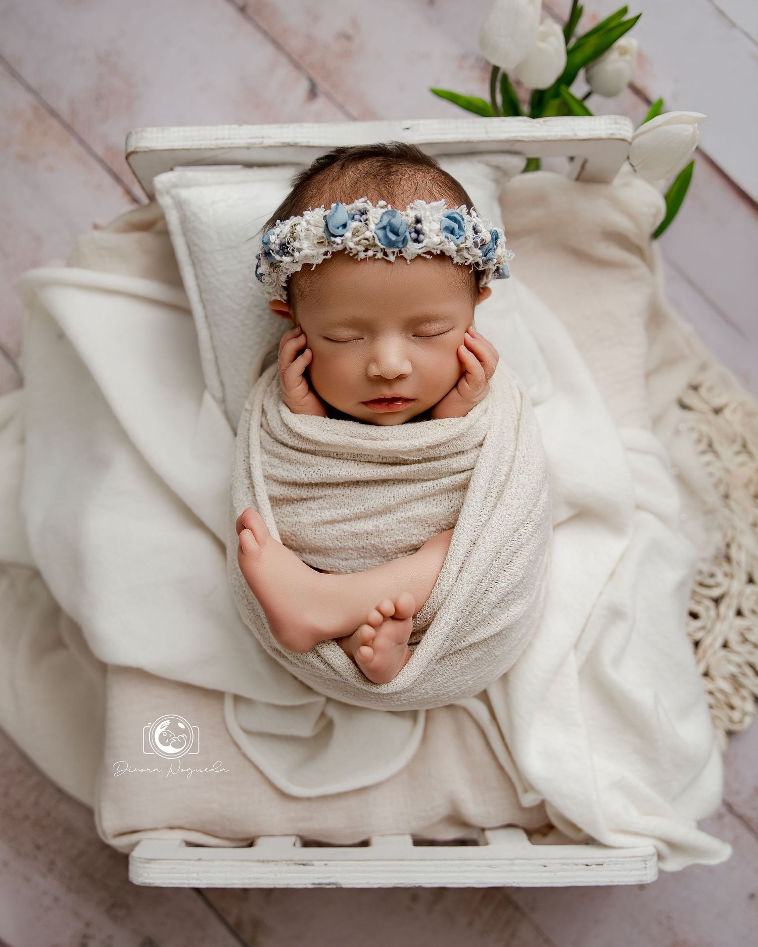 A sleeping newborn wrapped in soft cream fabric lies in a white wooden box wearing a delicate floral headband.