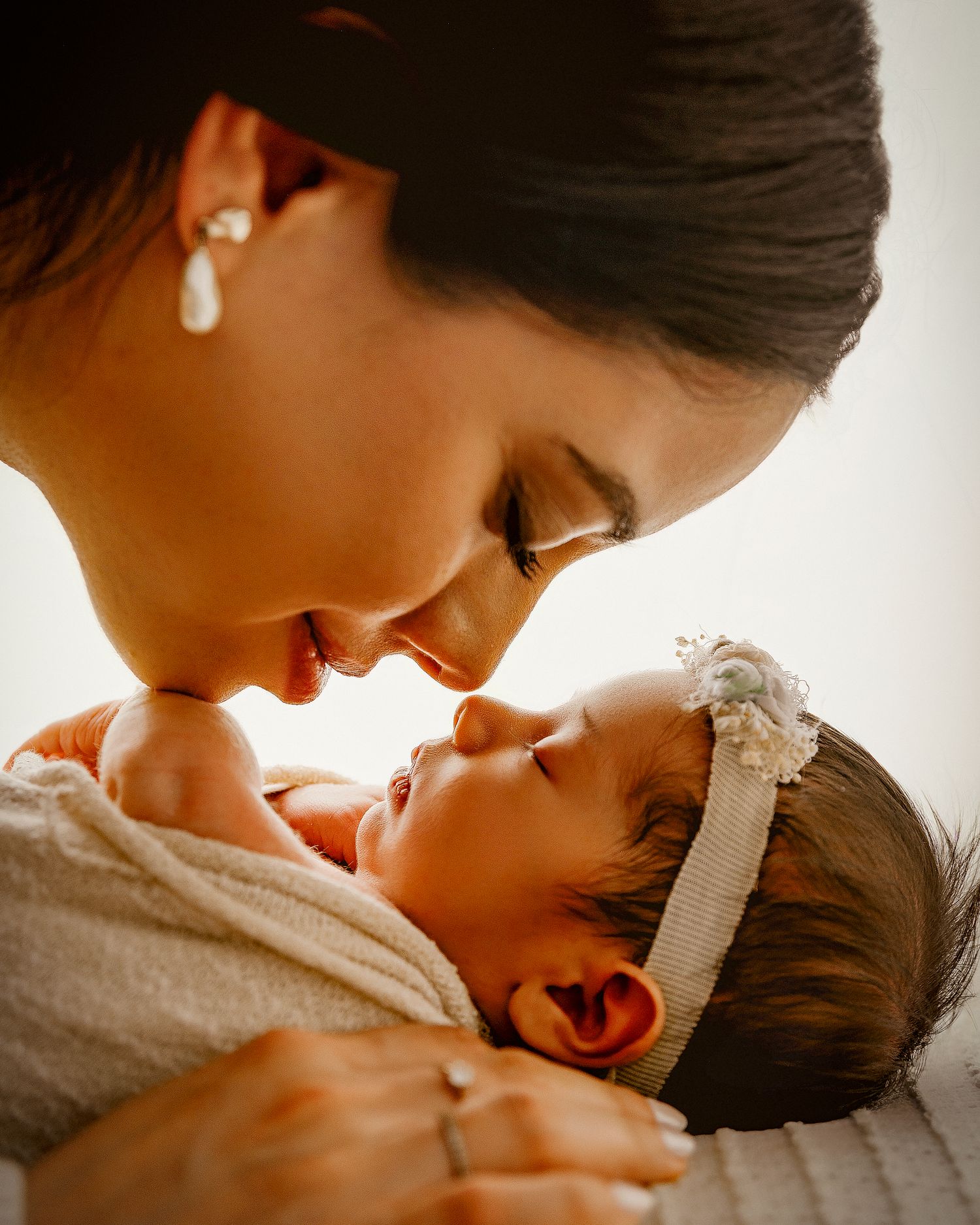 A tender moment as a dark-haired mother gazes lovingly at her newborn baby girl wearing a white flower headband.