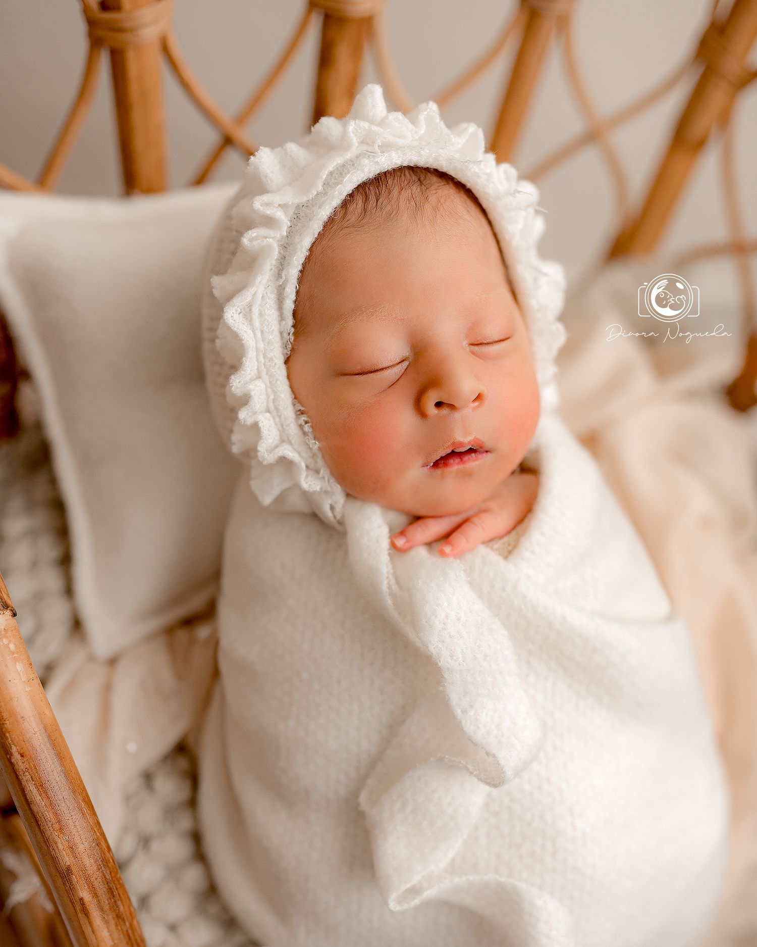 Sleeping newborn in white bonnet and wrap resting in wooden chair with cozy cushions.