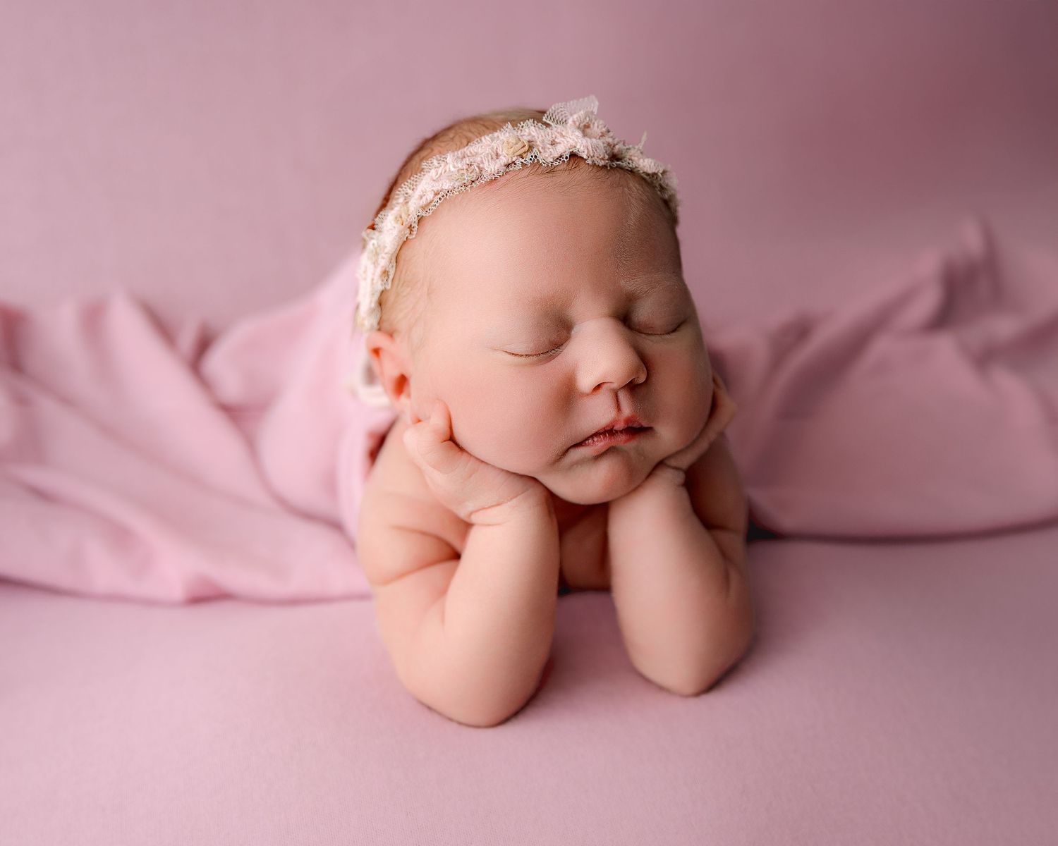 Newborn in delicate floral headband sleeping on pink backdrop with hands under chin.