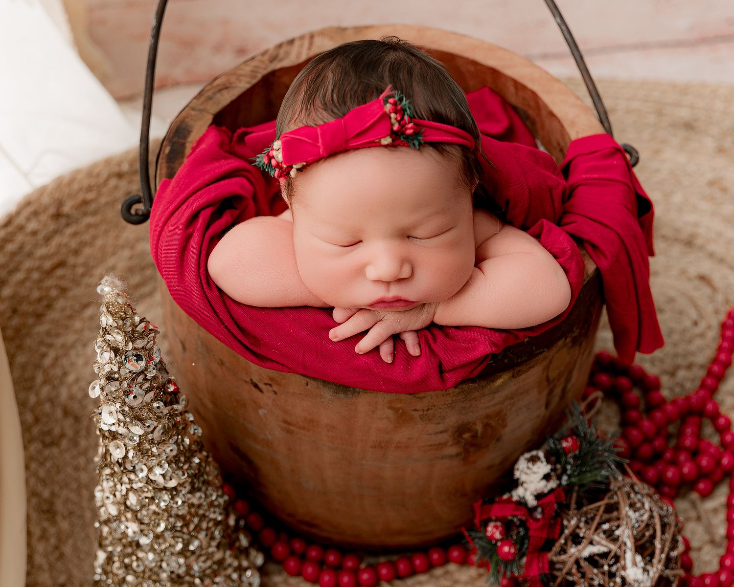 Sleeping newborn baby in red wrap and bow headband posed in a wooden bucket with holiday decor.