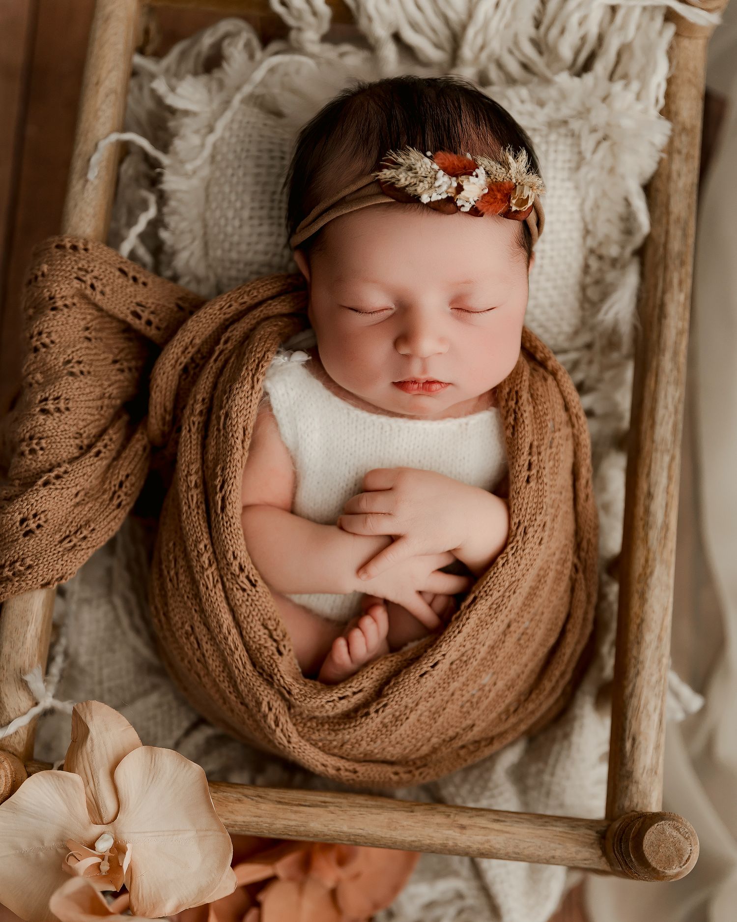 Newborn baby girl sleeping peacefully wrapped in brown knit, wearing floral headband in a rustic wooden bed prop.