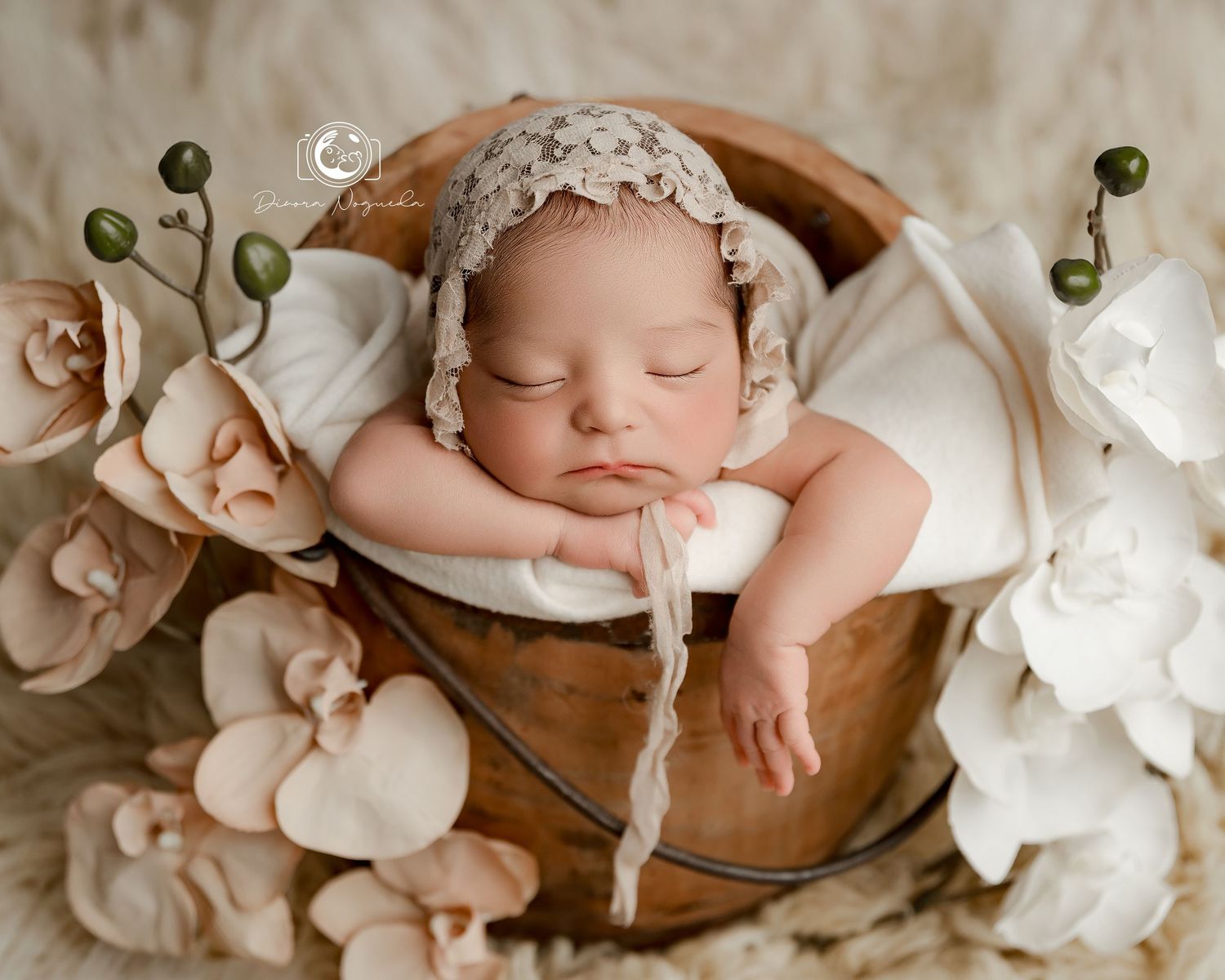 Sleeping newborn wearing delicate lace bonnet nestled in wooden bowl surrounded by pink and white roses.