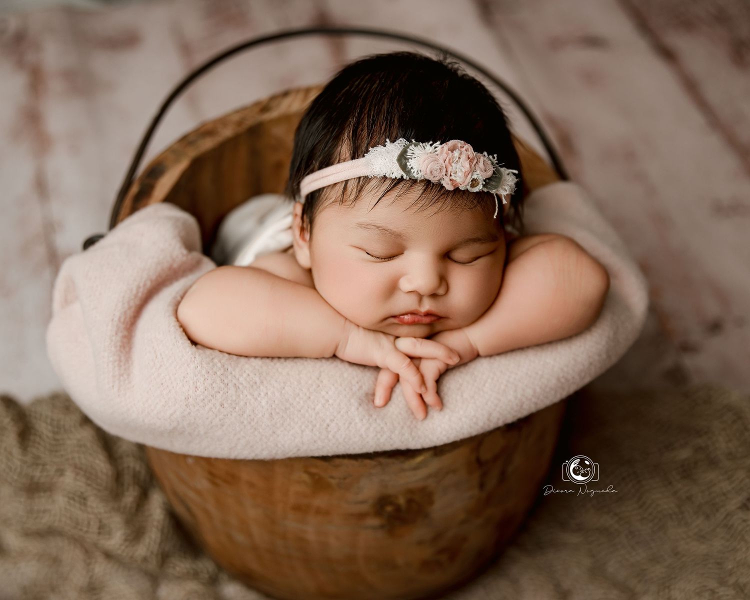 Sleeping newborn baby wearing delicate headband rests peacefully in wooden bowl lined with soft beige fabric.