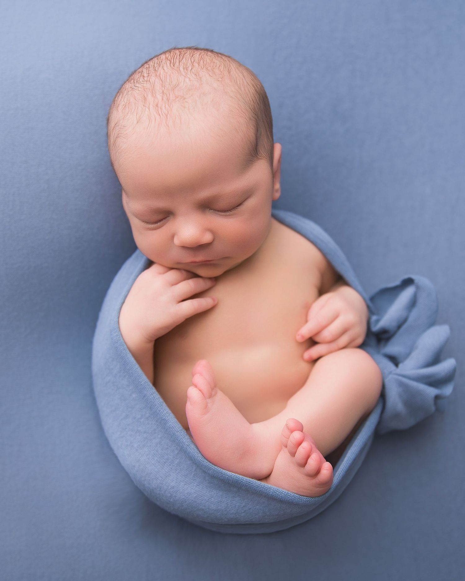 Sleeping newborn baby curled up on soft blue blanket in professional studio portrait setting.