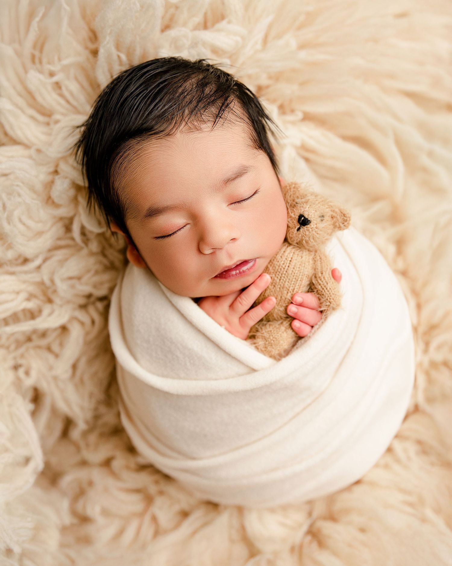 Sleeping newborn wrapped in white swaddle holding small teddy bear on fuzzy cream backdrop.