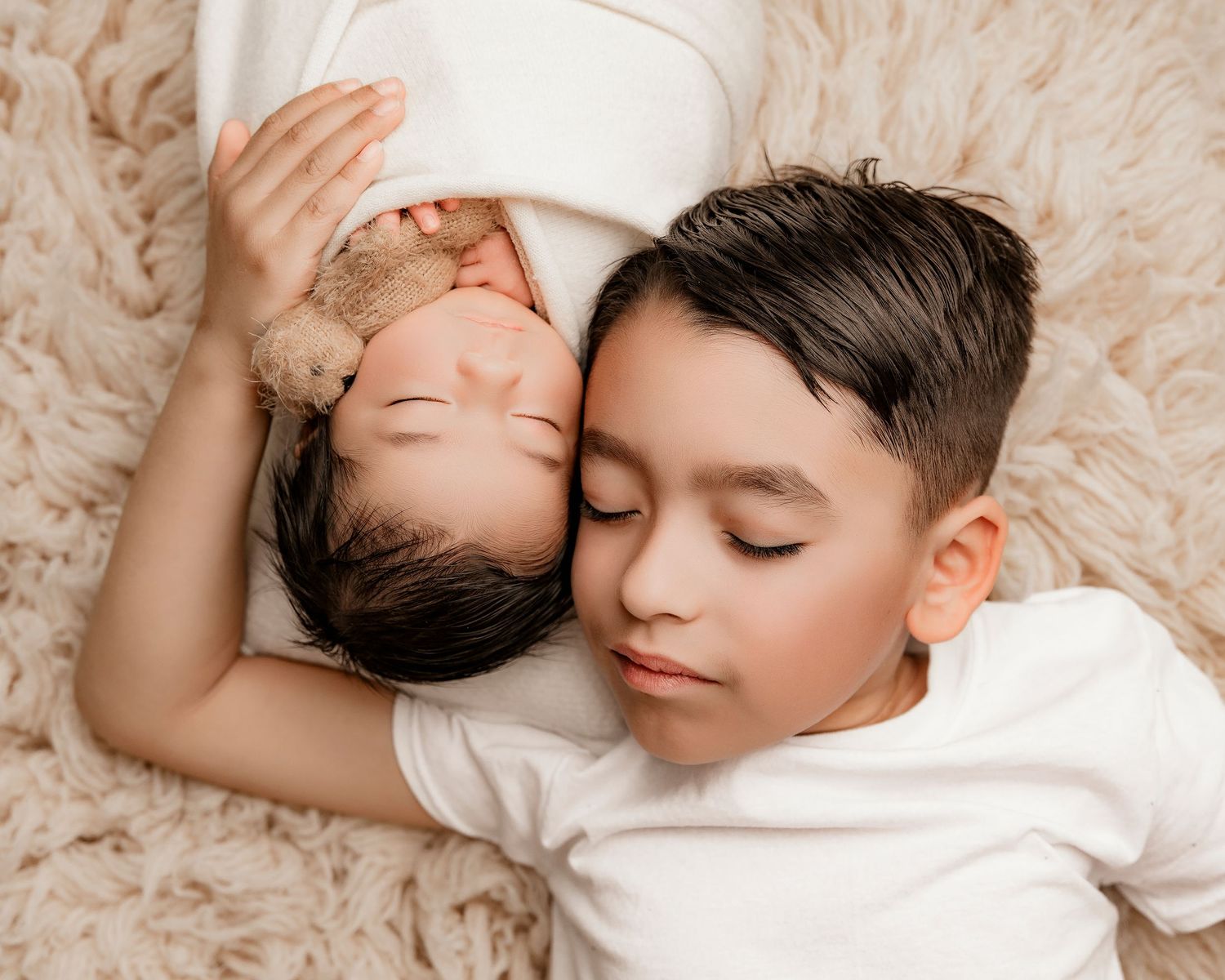 Two children resting together on a cream-colored textured blanket during a portrait session.