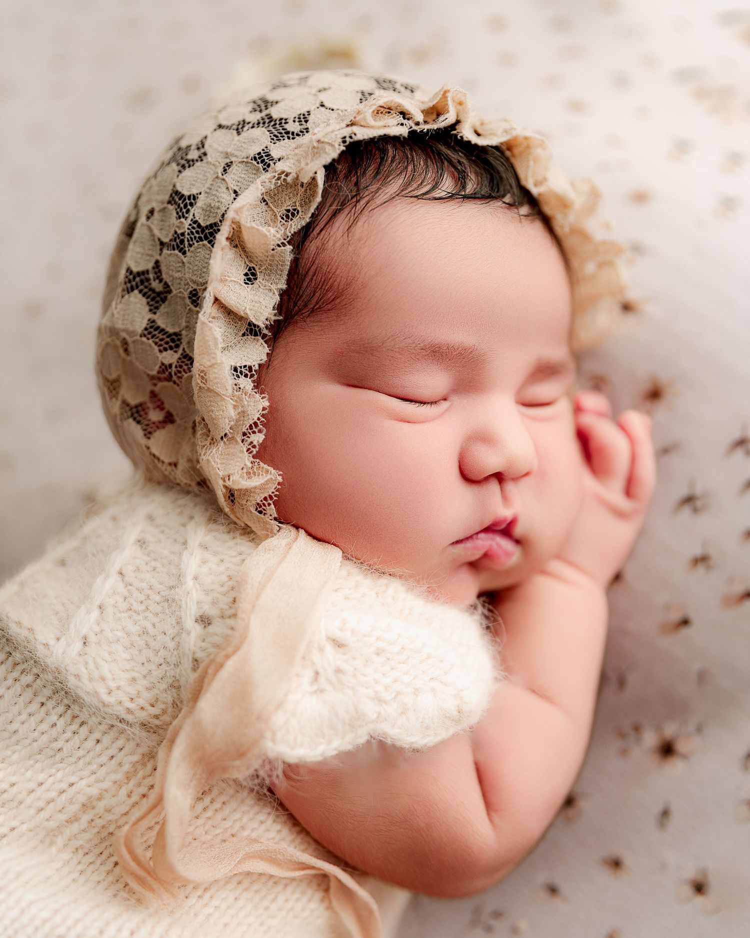 Sleeping newborn wearing vintage style bonnet and lace outfit on textured backdrop.