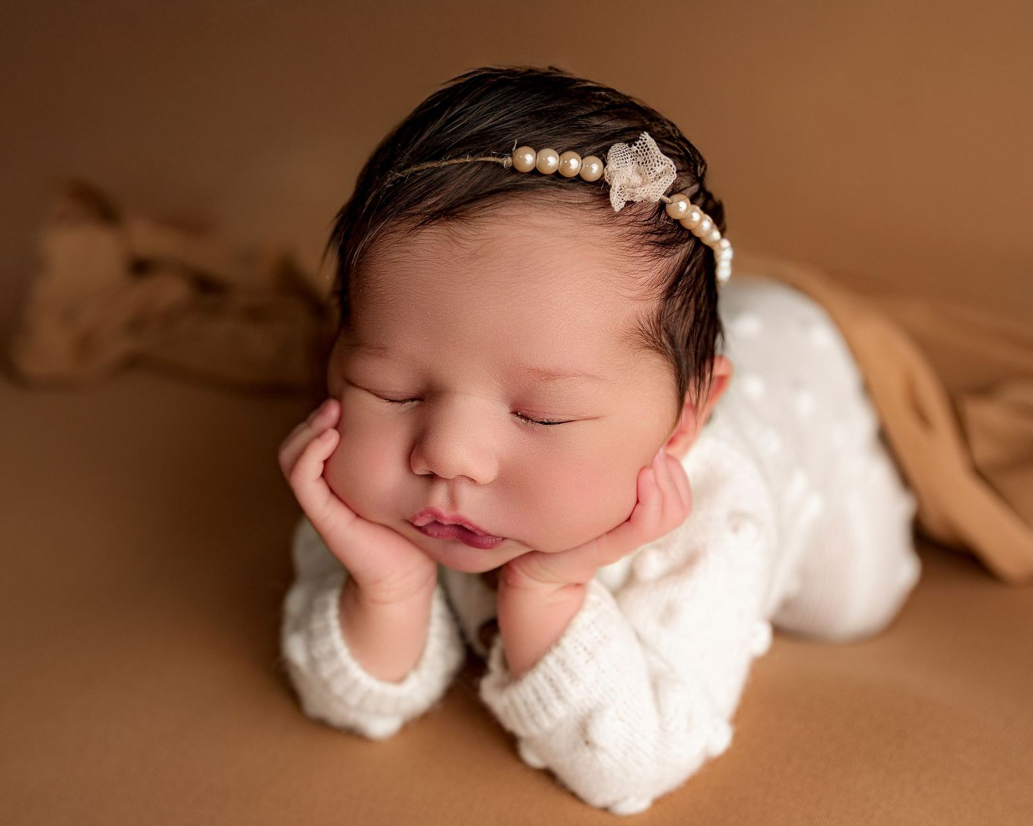 Sleeping newborn in white outfit poses with hand under chin on brown backdrop.