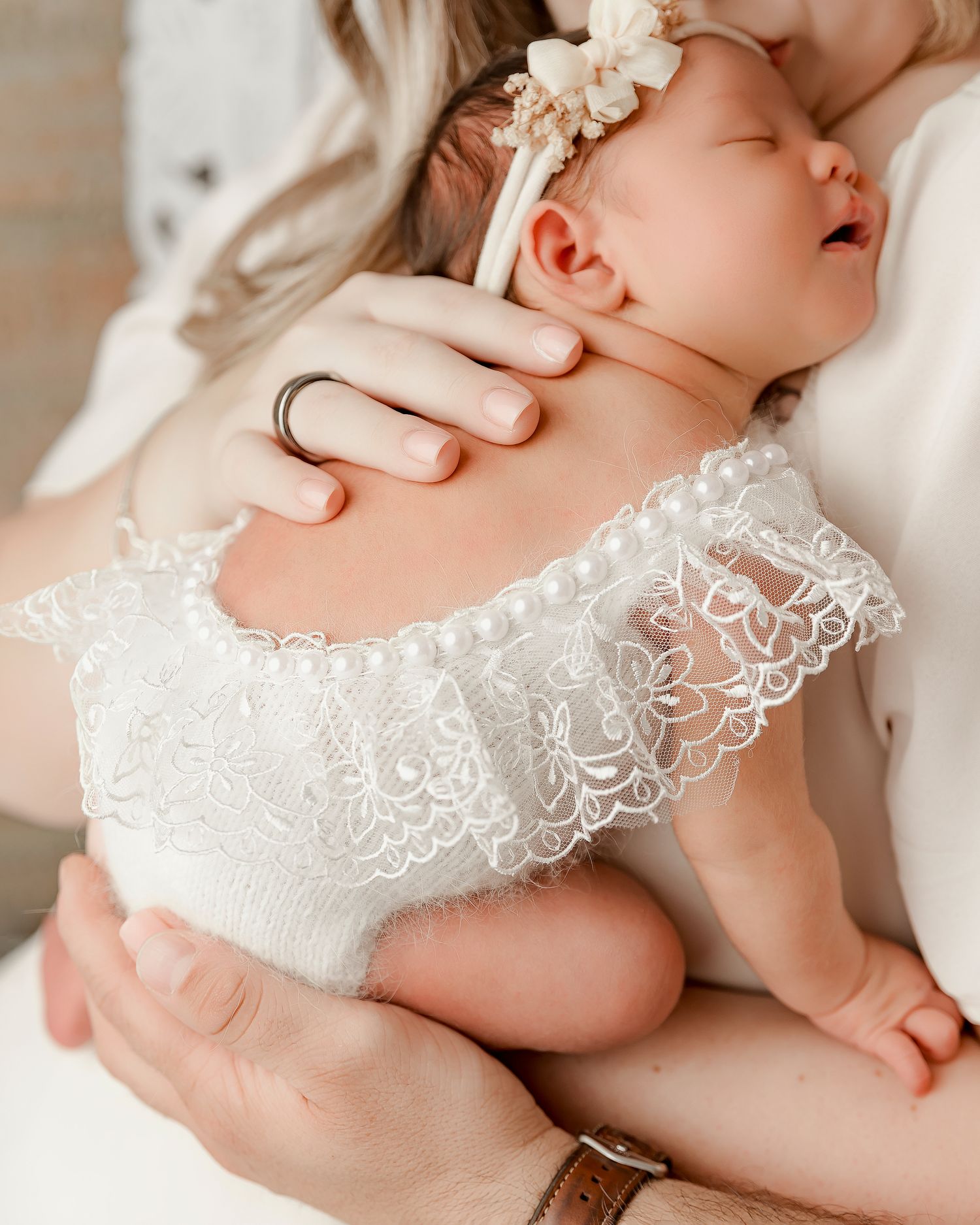 Newborn baby in white lace outfit and floral headband cradled in mother's arms during professional photo session.