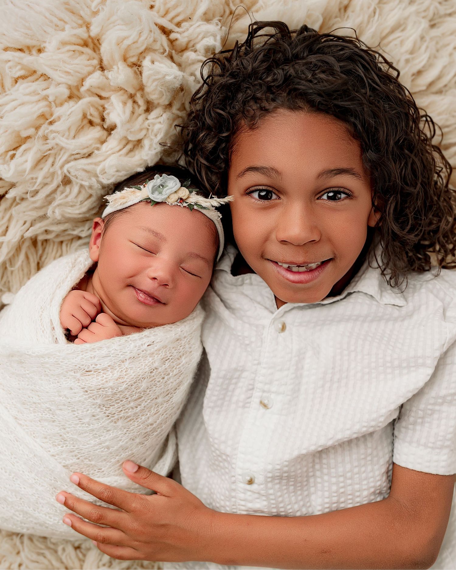 Smiling newborn swaddled in white with floral headband beside older sibling on cream textured blanket.
