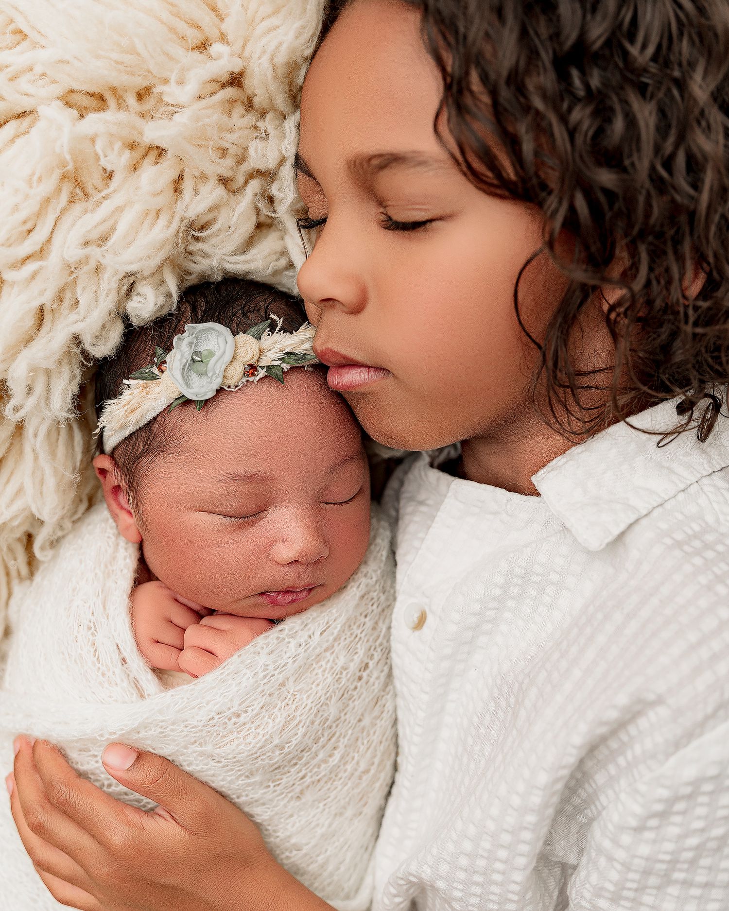 Older child gently kissing sleeping newborn baby wrapped in white swaddle with floral headband.