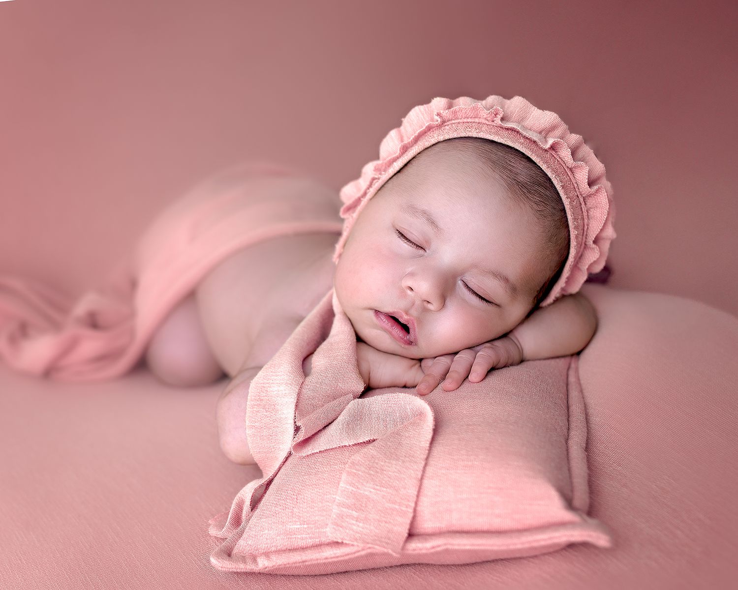 Peaceful sleeping newborn in pink bonnet posed on soft pink backdrop.
