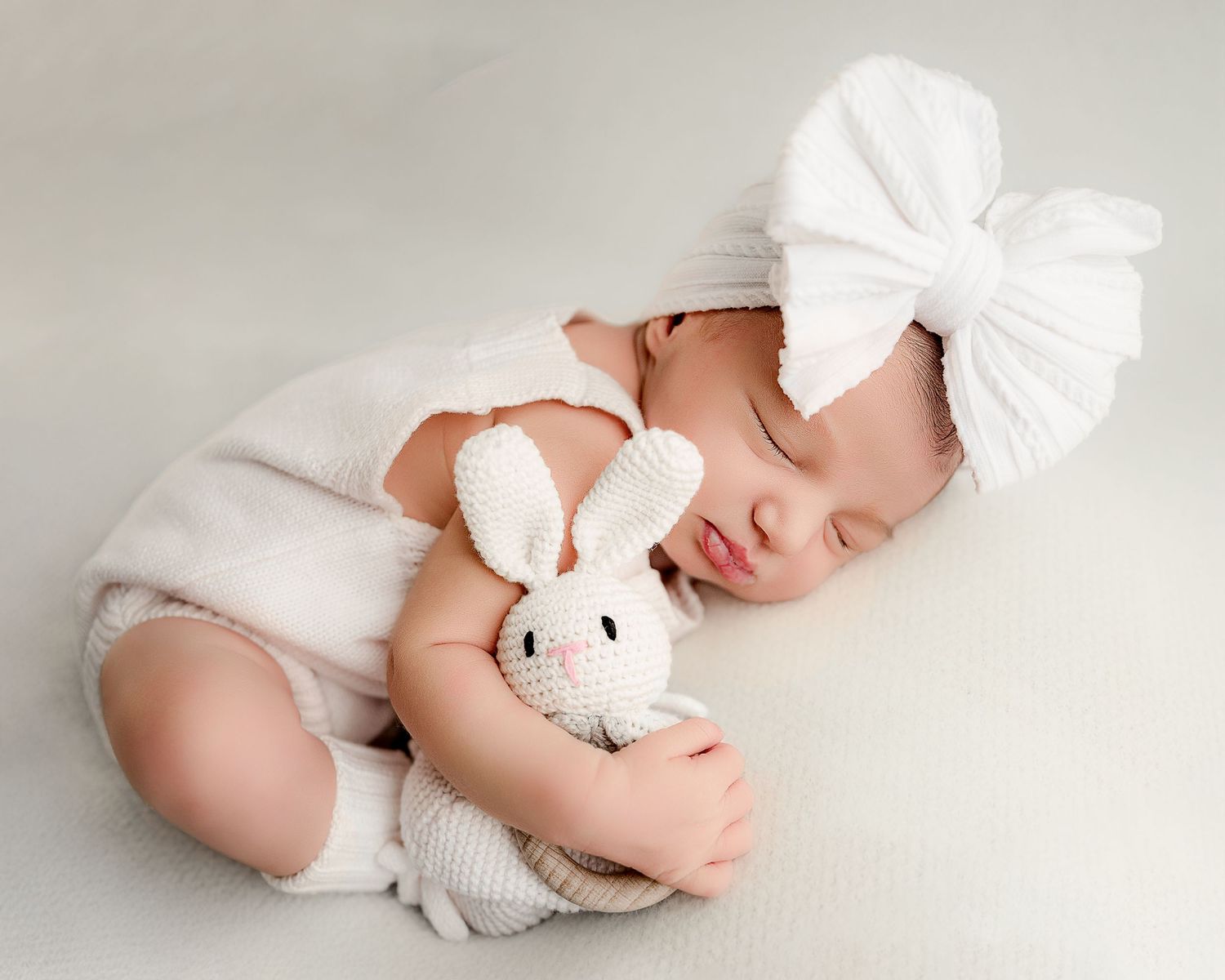 Sleeping newborn in white outfit with bow headband cuddling small bunny toy.