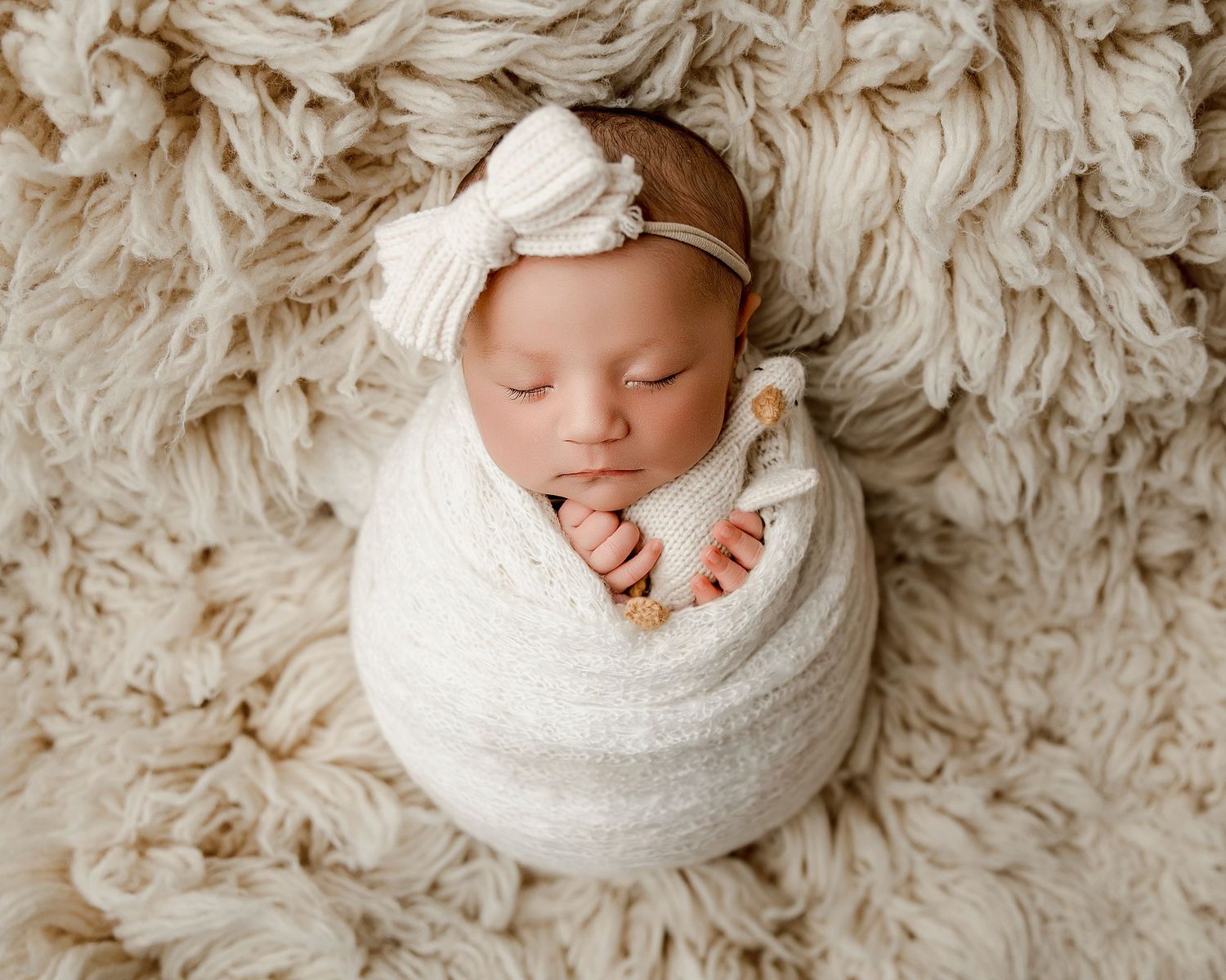 Newborn baby swaddled in white wrap with bow headband, sleeping peacefully on fluffy cream textured blanket.