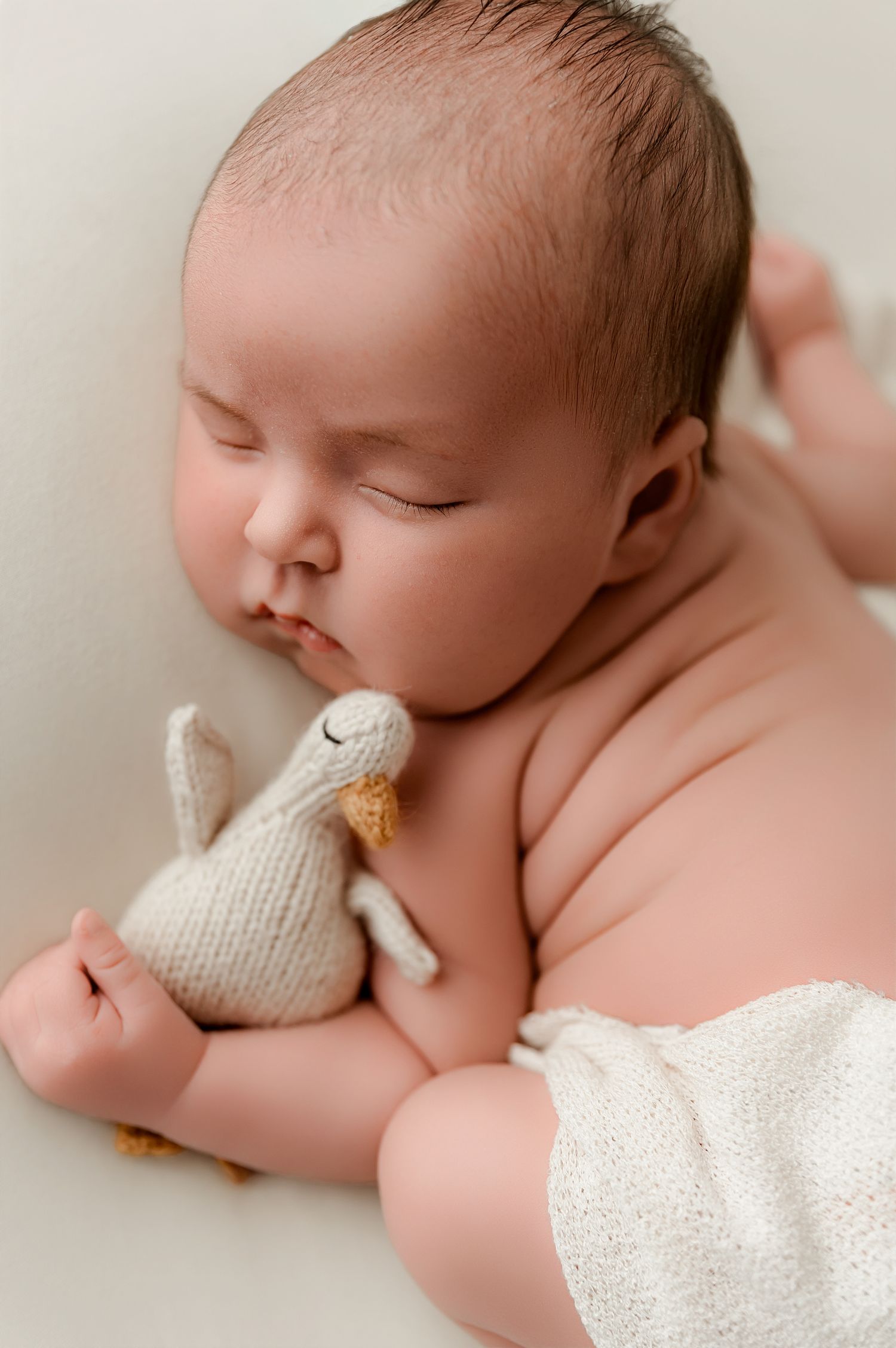 Peaceful newborn baby sleeping while holding a small knitted bunny toy, wrapped in a soft white blanket.