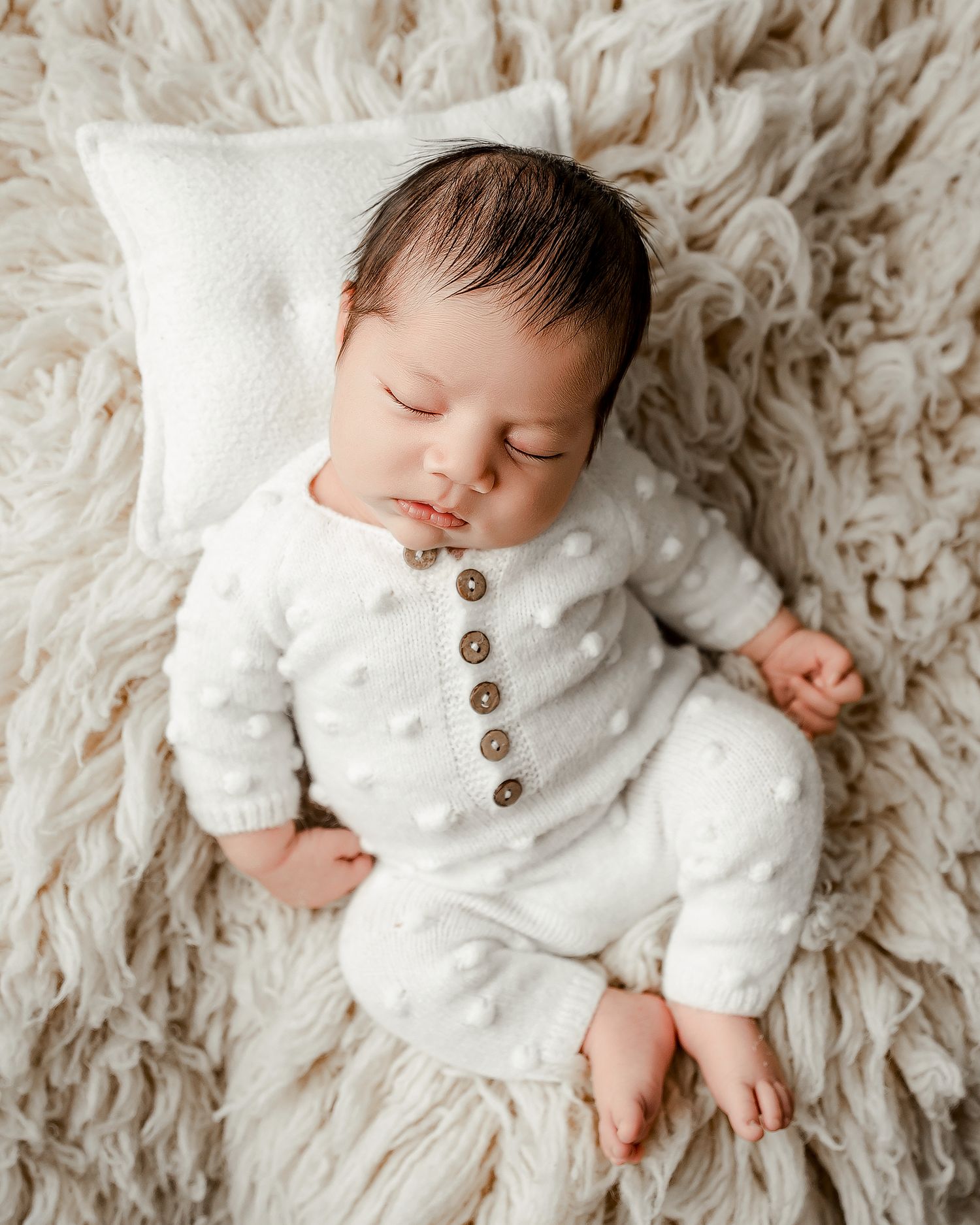 A sleeping newborn wearing a white button cardigan rests peacefully on a textured cream-colored blanket.